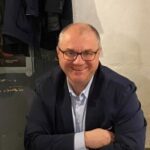 Adam Iwanow w okularach, ciemnej marynarce i jasnoniebieskiej koszuli, uśmiechnięty przed obiektywem kamery.