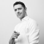 Krystian Cieślik na szarobiałym tle, ubrany w białą koszulę, wskazującego palcem na obiektyw aparatu.