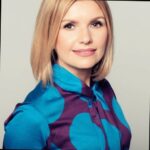 Sylwia Rogalska w krótkich do ramion blond włosach, ubrana w niebieską koszulę z okrągłymi fioletowymi elementami. Jej twarz skierowana jest w kamerę.