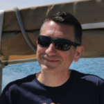 Krystian Cieślik na szarobiałym tle, ubrany w białą koszulę, wskazującego palcem na obiektyw aparatu.