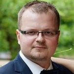 Krystian Cieślik na szarobiałym tle, ubrany w białą koszulę, wskazującego palcem na obiektyw aparatu.