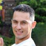 Krystian Cieślik na szarobiałym tle, ubrany w białą koszulę, wskazującego palcem na obiektyw aparatu.